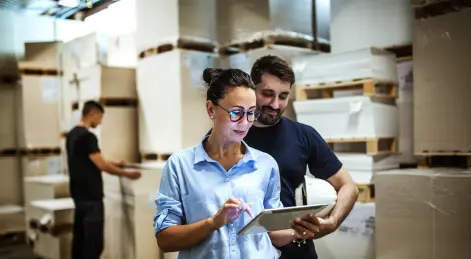 imagen mujer con lentes mostrando inventario a hombre con barba de fondo una bodega con cajas